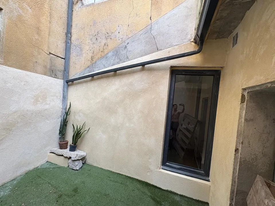 Façade crépie avec sa baie vitrée noire, le gazon synthétique et une plante en pot.