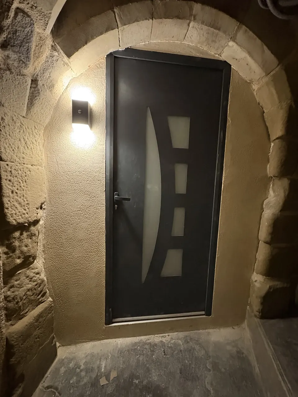 Nouvelle porte anthracite à vitrage galbé, éclairée par une applique murale.