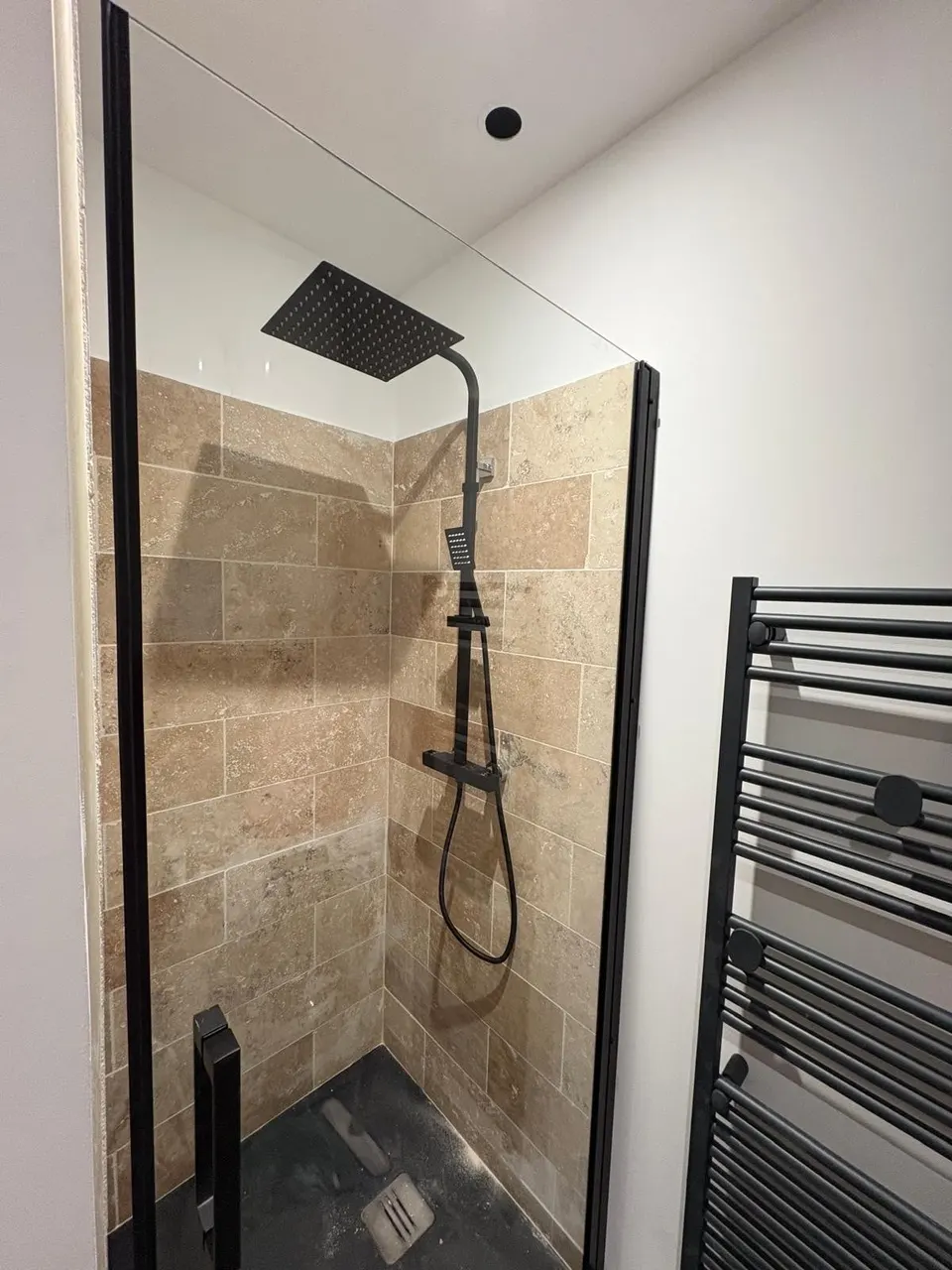 Douche terminée : travertin, robinetterie noire et paroi vitrée